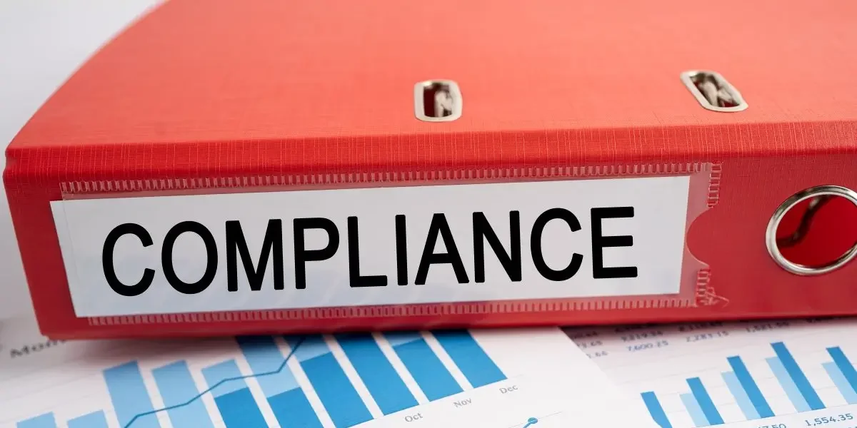 A red binder labeled compliance sits on financial charts and graphs, representing the importance of payroll compliance in business.
